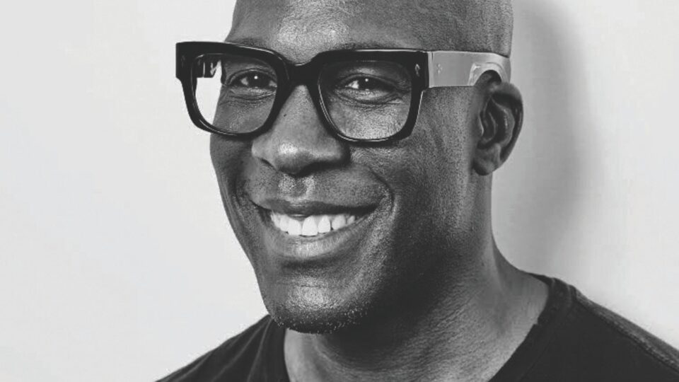 A black-and-white portrait of a bald man wearing large, thick-rimmed glasses and a dark shirt. He is smiling warmly and looking slightly off to the side. The background is plain and light, highlighting the subject's face.