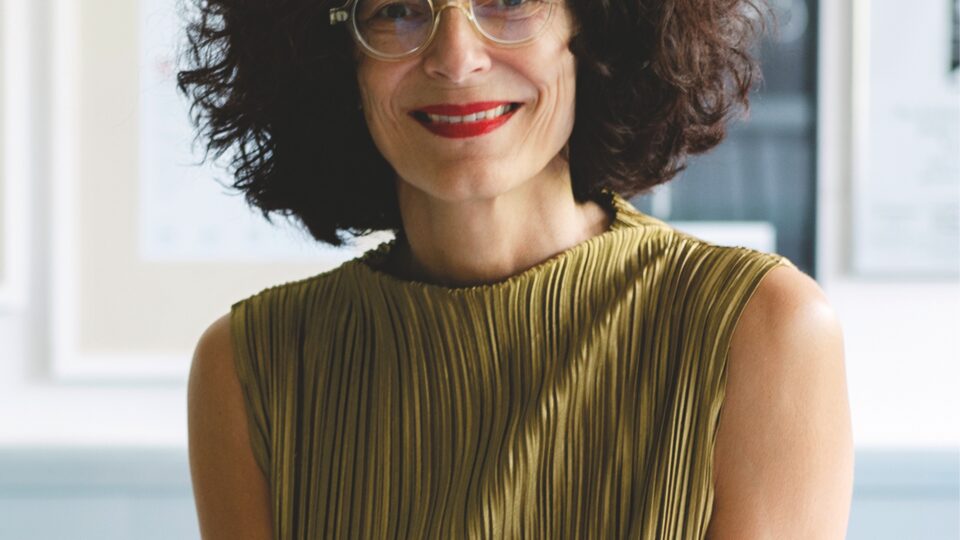 A portrait of Shirin Frangoul-Brückner smiling warmly at the camera. She is wearing a pleated, olive-green sleeveless top, with curly dark hair and round glasses. The background is softly blurred, with framed works visible behind her.