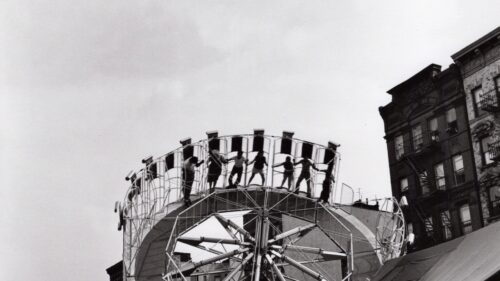 Black and white photograph by Arthur King showing a spinning carnival ride (Round-Up) in front of historic urban buildings, featured in the Weathered exhibit.