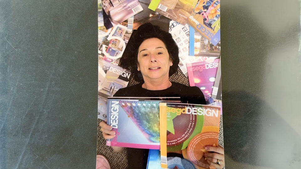 Leslie Dilworth lying on the floor surrounded by SEGD Design magazines, holding two issues and smiling at the camera, representing her leadership and impact in environmental graphic design.